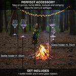 Outdoor setting with bottle and wine glass holders near a campfire, surrounded by trees.